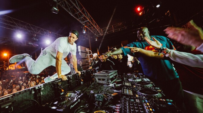 NFL player Rob Gronkowski does a jump off dance move from Shaquille O'Neal's DJ turntable setup. Shaq is standing on the other side of the tables.