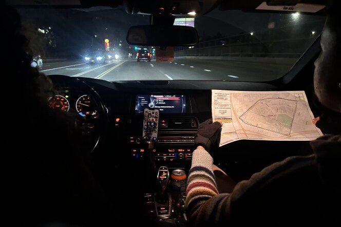 The interior of a car from the vantage point of the backseat. The person on the right in the passenger seat is holding up a printed map with both hands, which are wearing fingerless black gloves. The interior is slightly illuminated by buttons on the dashboard, with streetlights and car headlights shining through the front windshield. 