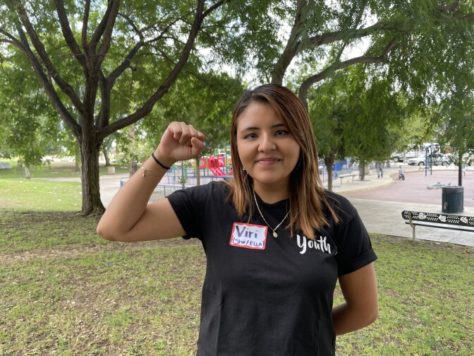 A young woman with shoulder-length hair holds up her right fist while smiling at the camera. There is a playground with a few children behind her, along with trees and grass. 