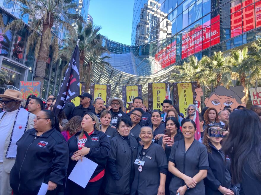 A crowd of people are standing before a large hotel in downtown Los Angeles. There are several signs and a flag as  well as palm trees lined behind them with a blue sky peaking behind them. The front row shows five women smiling. 