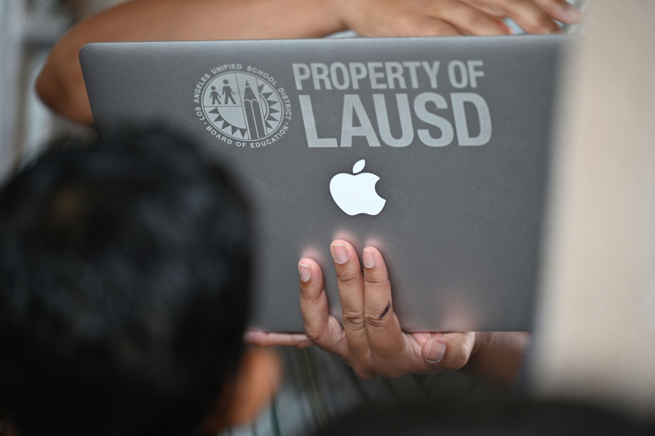 A hand with medium skin tone holds up a silver laptop with the image of an Apple and the text LAUSD on the back of it.