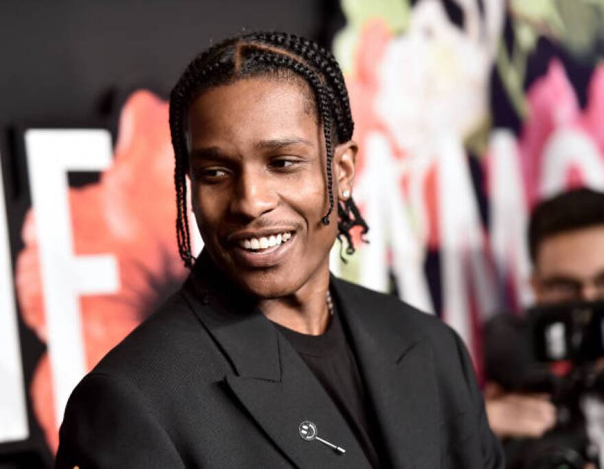 Rapper ASAP Rocky, whose hair is braided and is wearing a black suit with a black suit, smiles at an event.
