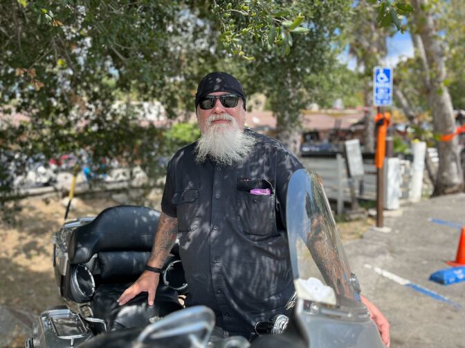 A man with a long white beard and moustache, sunglasses, a black short-sleeved shirt and a black band tied around his head poses alongside his motorcycle outside Cook's Corner in Trabuco Canyon. 