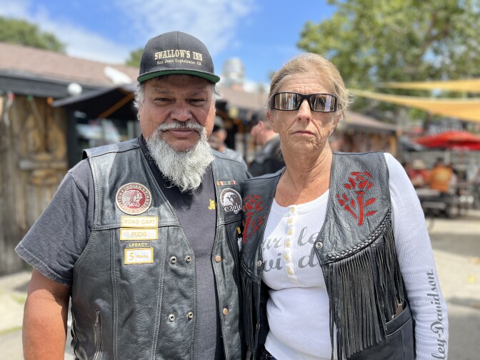 Two people, one wearing a black motorcycle vest, the other wearing a black leather fringe vest over a white long-sleeved shirt with a Harley-Davidson logo on it, solemnly pause outside Cook's Corner. 