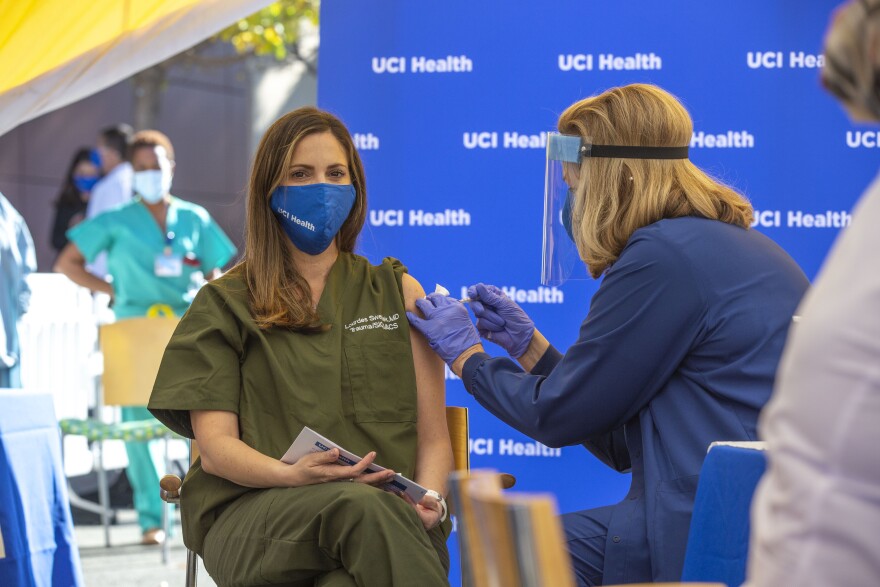 A seated woman wearing green medical scrubs and a blue mask receives a coronavirus vaccine shot.