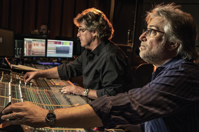 Two male presenting people in front of a console that looks like an audio mixing board.