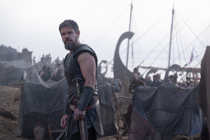 Actor Matt Damon stands on a gloomy shore wearing dark Greek armor. Tarps and historically Greek boats can be seen in the background.