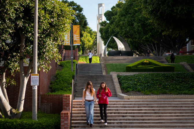 Two femme presenting students walk down university stairs outside. There are trees and greenery