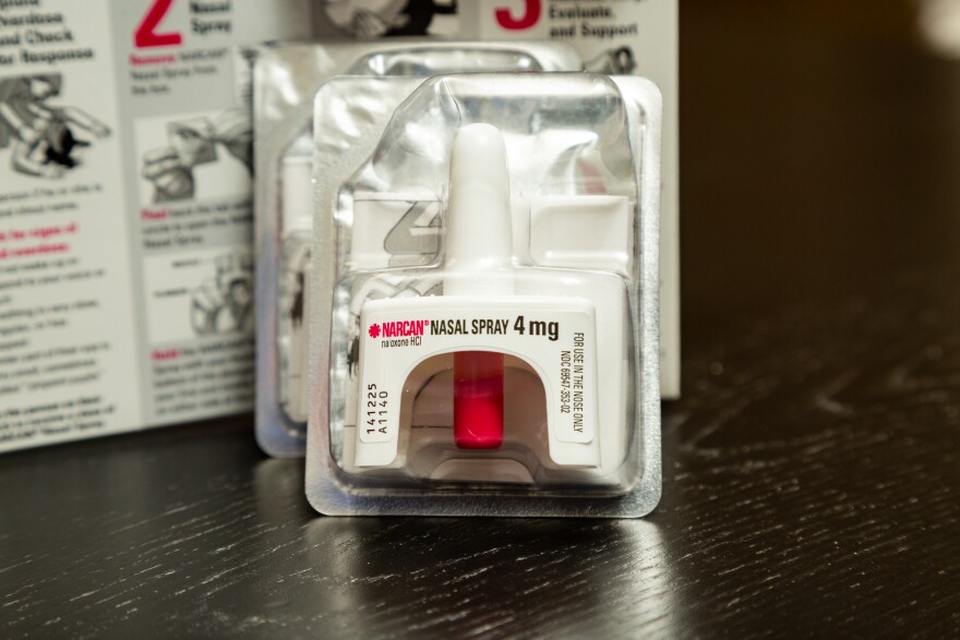 A small plastic and foil package contains a plastic device with a protrusion shaped to fit in the nose. The device is red and white and has lettering that reads "Narcan Nasal Spray 4 mg."