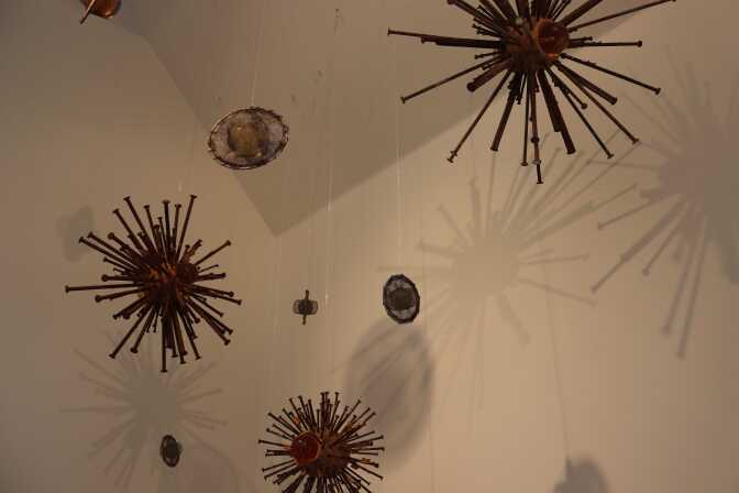 Several small sculptural objects are hung from a ceiling. They are spherical. Some have many stick-like pointers coming out from them, and others look like circles with smaller circles inside. 