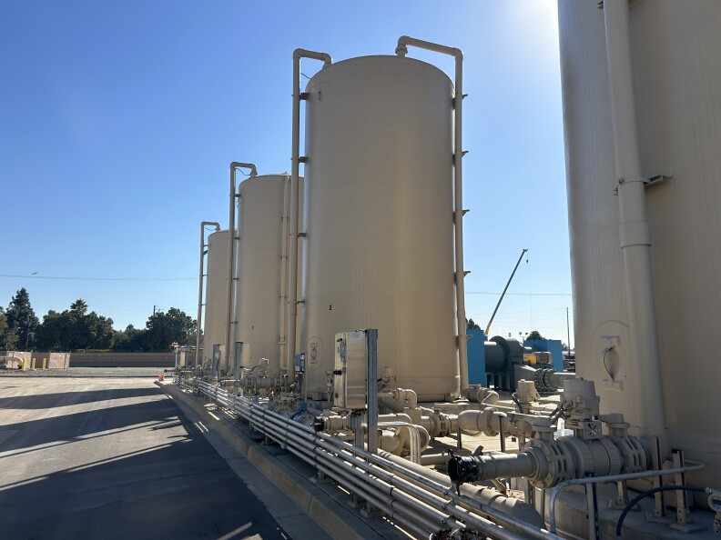 Four large cylindrical tan tanks in a row under sunny blue skies in a cement driving area. 