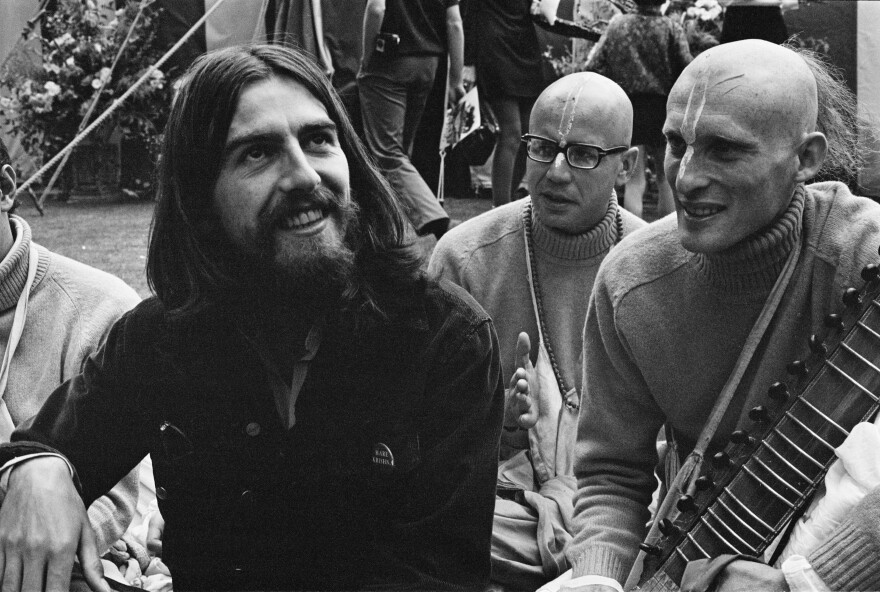 George Harrison, with long shoulder-length hair and a beard, and wearing a dark jacket with a pin on it, smiles as he looks up and off to the side. Two men behind him with their faces painted look toward him — one of them holds a sitar.