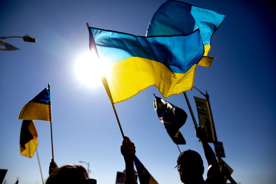A large half light blue on top, yellow on bottom Ukrainian flag is waved by a hand in shadow in front of a dark blue sky in front of the sun. Two other hands wave smaller Ukrainian flags in the air next to an obscured dark street light lamp post during the daytime in Santan Monica.