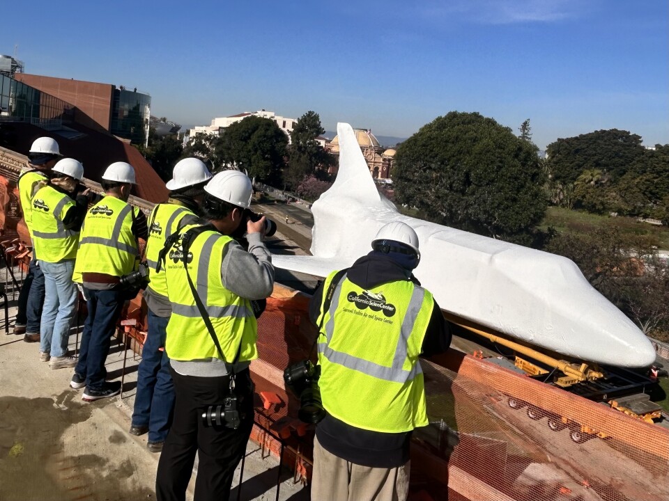 Six people wearing neon green safety vests and white hardhats are looking over the ledge of a multi-story building towards a massive, stark white space shuttle orbiter.
