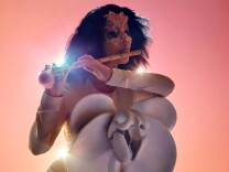 Björk, lit from behind and wearing a gold dress, with a golden tiara, and a series of white shapes in front of her, plays a flute.