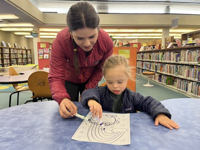 A woman with a ponytail and cranberry jacket helps a 3-year-old boy draw.