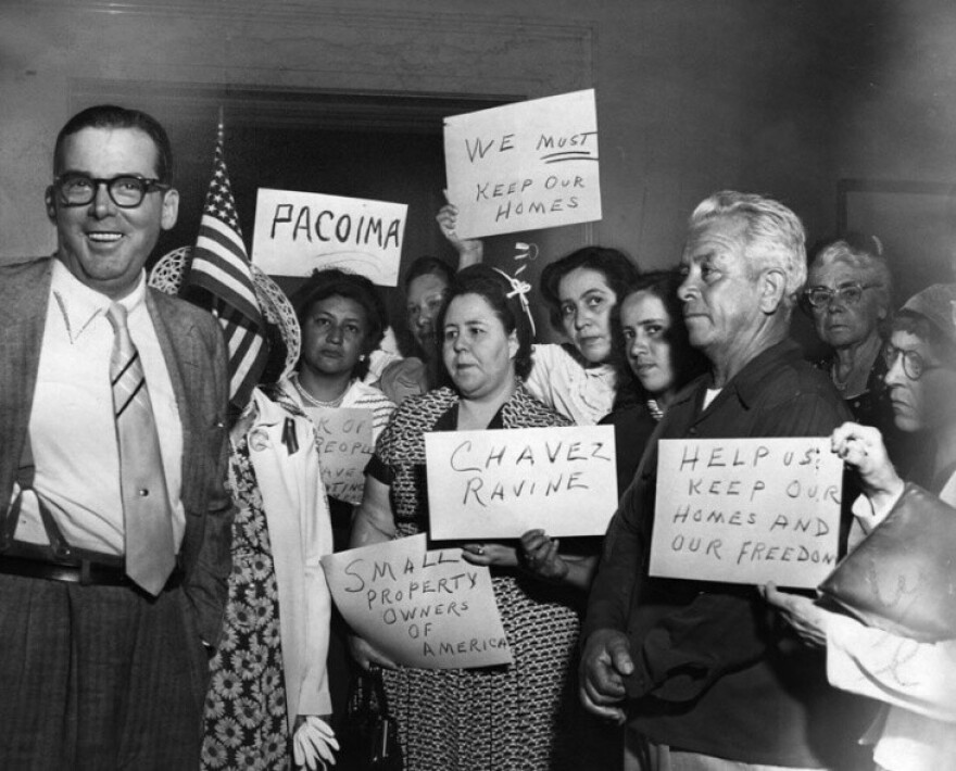 In a black and white photo, multiple Chavez Ravine residents , most middle aged and some elderly, hold up paper signs protesting the displacements. Some signs say “We Must Keep Our Homes” and “Small Property Owners of America”. They stand in a crowd towards the right, next to a portly man in thick black glasses and slicked back hair. He cheery demeanor is juxtaposed by the angry and concerned faces in the crowd.in between the crowd and the nan is an American flag.