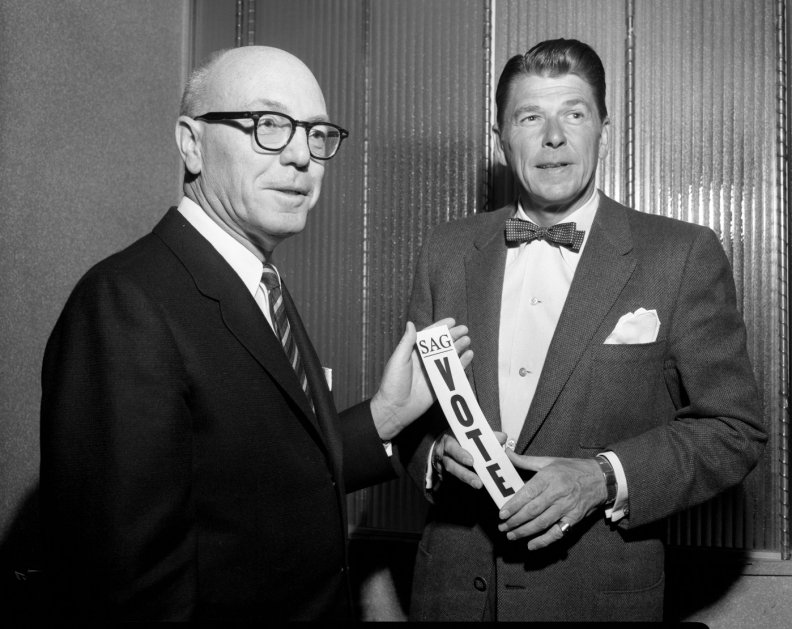A black and white photo shows two light-skinned men in dark suits from 1960; one is bald with glasses, the other (Ronald Reagan) has dark hair and is wearing a bow tie. They are holding a piece of paper which says "SAG VOTE".