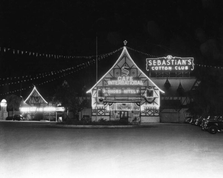 a building with a pointed roof outlined in glowing lights has a glowing sign that reads "Cafe International." a sign behind the building reads "Sebastian's Cotton Club."