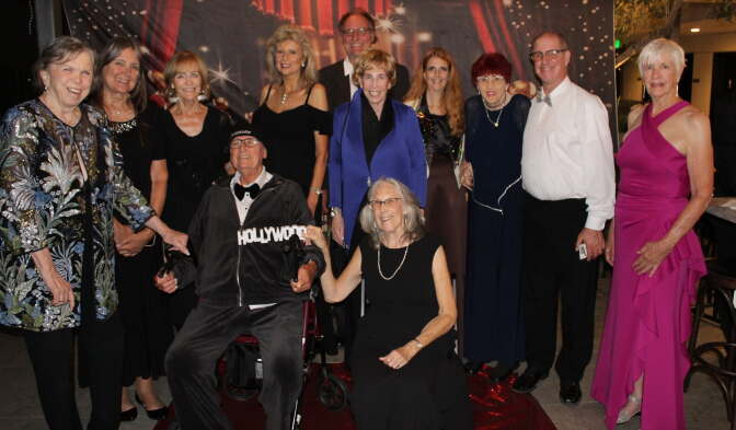 A group poses in formal attire on a red carpet before a Hollywood-themed backdrop, one person wears a tracksuit with a small 'Hollywood' sign held in front of it.