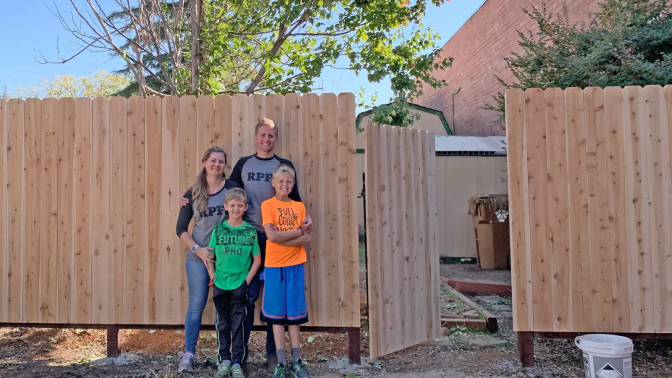 A family of four — a man, a woman and two boys, all with light skin, blond hair and blue eyes — stands in front of a newly built wooden fence
