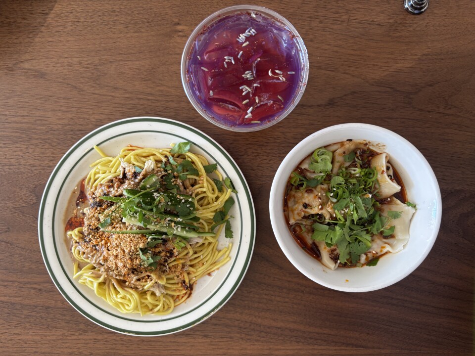 Overhead view of two dishes on wooden table: yellow sesame cold noodles topped with ground peanuts, cucumber and fresh herbs on left; white bowl of chili wontons in red chili oil topped with scallions and cilantro on right; purple Taiwanese fruit tea in glass above.