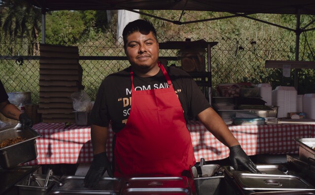 A medium dark Latino man with a black shirt, red apron and black latex gloves standing in front of a food station. In the background, there are food containers and materials.
