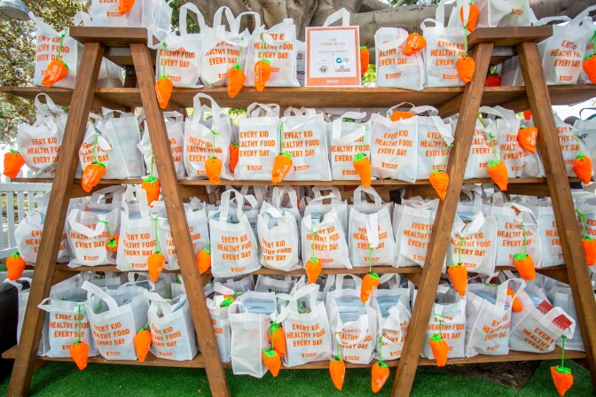 Image is showing a 4 tier shelf and on every shelf is packed with a white goodies bag that says 'Every kid, healthy food, every day. The bags also have an origami shaped carrot tied around the handles.