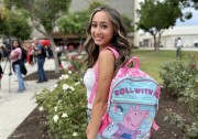 A girl with medium-light skin tone turns for a portrait. On her back is a backpack that says "Roll with it," and has a picture of Peppa Pig, a bright pink cartoon pig.