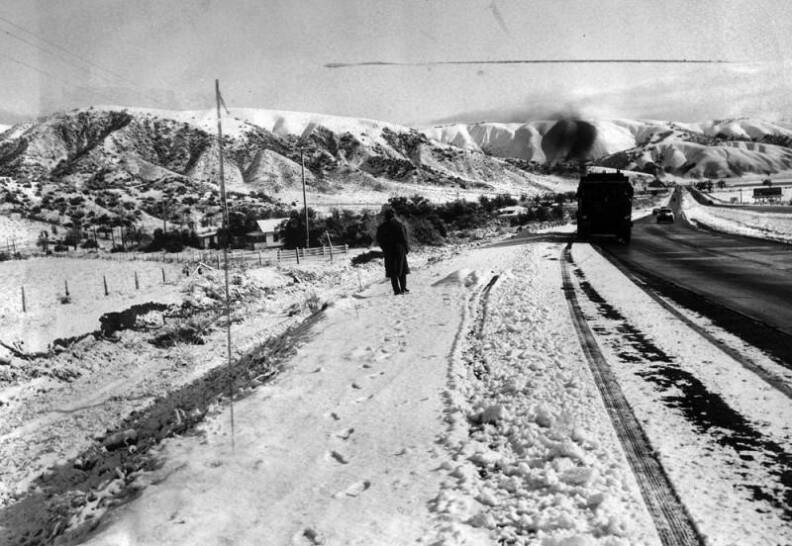 A figure walks through snow on the side of a road as vehicles drive by. Snow-covered mountains line the background.