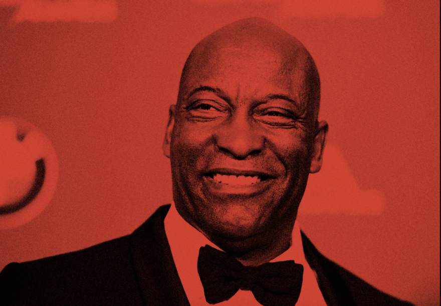 In this red-filtered image, a Black man in a suit smiles widely at something off camera as he attends an awards show.