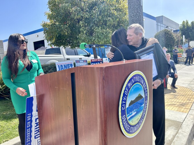 A woman in green stands to the left of a podium with the seal of the City of Huntington Beach on it. On the right side of the podium, a woman in black hugs an older man carrying a framed certificate. 