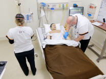 A man in a white uniform, bends over a hospital bed and examines a mannequin's arm. A woman stands next to the bed with her back to the camera. Her white uniform has the letters "RCC" on it.
