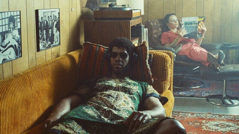 A Black man with headphones and a green African-styled outfit lays on an orange couch, black and white photos on the brown paneled wall next to him, including a photo of Bruce Lee. Behind him is a character played by actress Sarah Ramos on another chair, a record player in between them.