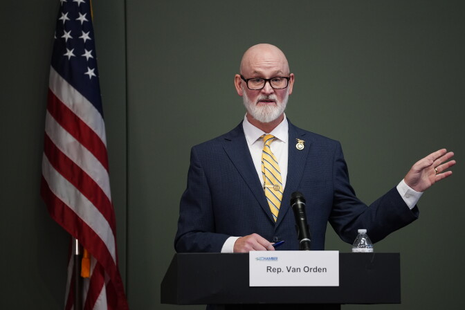 A bald man with a white beard and a light skin tone wears a blue blazer, a white shirt and a yellow and blue striped necktie. He gestures at a podium. An American flag is on the left.
