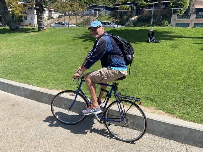 A man wearing shorts, a blue jacket, a black backpack and a light blue baseball cap rides a bicycle on a path beside a lawn in a park.
