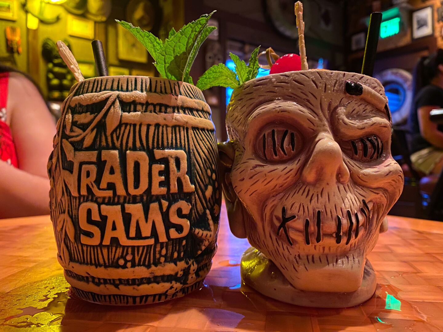 two tiki mugs sit on a wooden table. one is shaped like a barrel. one is shaped like a skull.