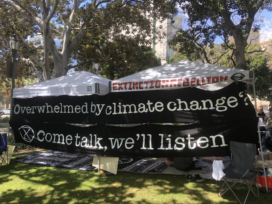 A black banner with white lettering that reads "Overwhelmed by climate change? Come talk, we'll listen" is stretched between two posts and mounted a grassy area of a plaza. Two pop-up tents, one labeled Extinction Rebellion, are behind the banner.