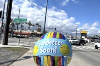 A small colorful balloon with yellow smiley faces and "feel better soon!" in blue-white text on the front is floating above a cement sidewalk near an intersection in the Los Angeles area.