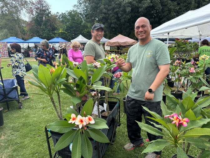 Um homem de pele clara e camiseta verde sorri em primeiro plano, ao lado de várias plantas de plumeria, enquanto outros compradores andam atrás dele.
