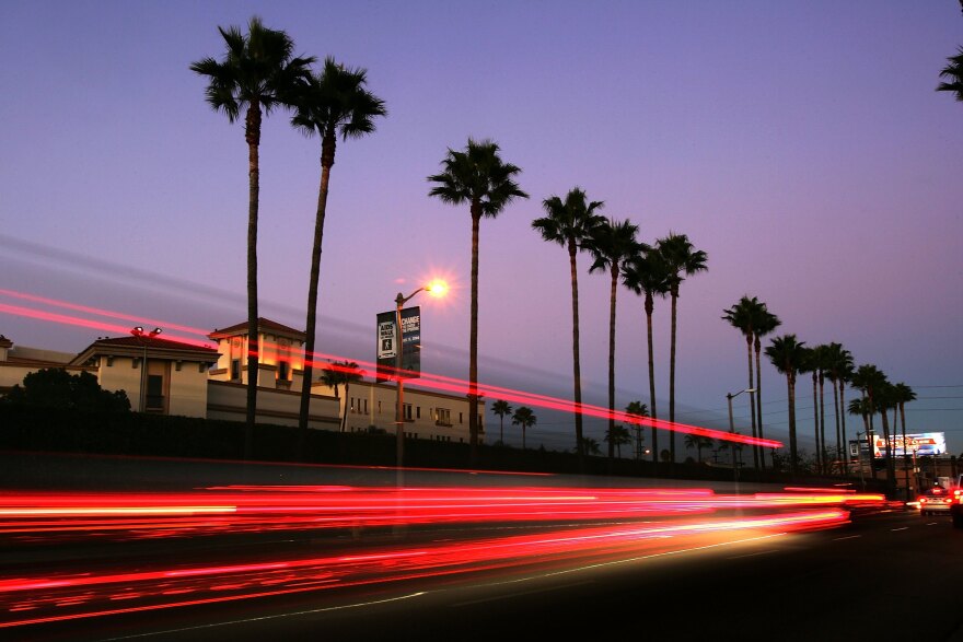 A line of towering palm trees with thin trunks are silhouetted along a busy street against a sky at sunset. The slow motion shot captures a streak of red tail lights on the street.