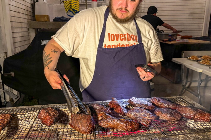 Chef Brad Thomas, wearing a navy apron over a Loverboy Tendencies t-shirt, uses tongs to tend to multiple hanger steaks on a charcoal grill in the backyard of a craftsman home at night, with a Weber grill visible in the background.