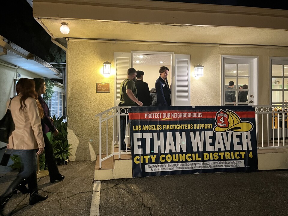 People walk down a well-lit pathway into a room and past a banner hanging on a metal railing that carries messaging in support of Ethan Weaver, a city council candidate.