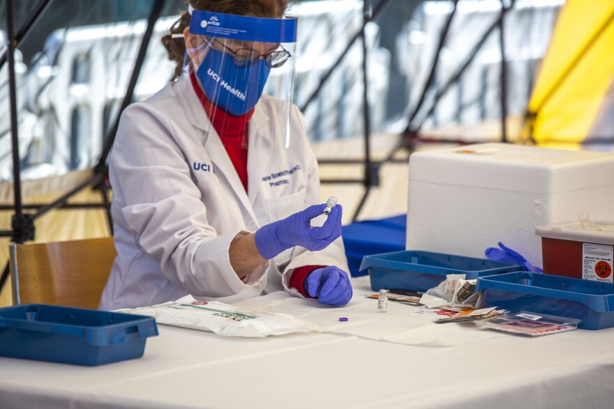 A medical professional wears a mask, face shield and rubber gloves as she inspects a vial of vaccine.