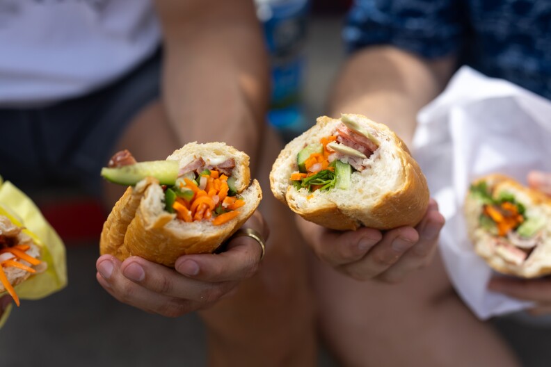 Two light-skinned hands hold baguettes filled with shredded carrots, cucumber, and a reddish meat item.