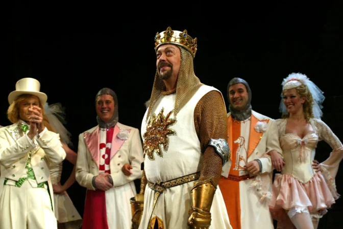 Tim Curry, wearing a costume of a king, looks up out of frame as other performers stand on stage behind him.
