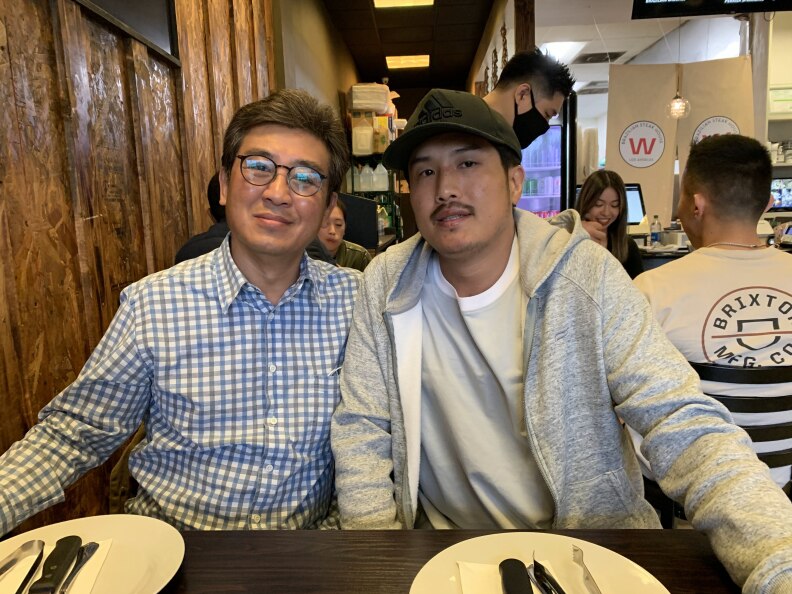 A middle-aged Asian man with medium-light skin tone wearing a patterned button up and glasses sits a restaurant table next to an Asian man in his 30s or 40s wearing a gray shirt and gray sweatshirt and Adidas baseball cap.