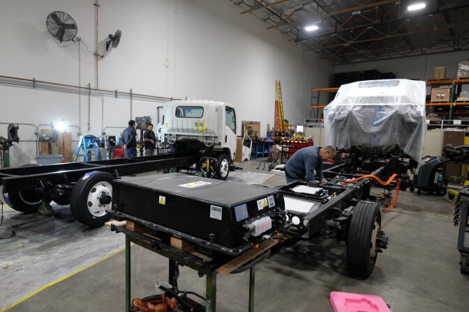 People work on two Isuzu trucks in a large warehouse. A lithium-ion battery that will be installed in one of the trucks is in the foreground. 