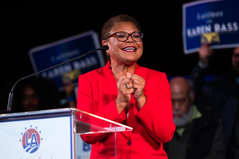 Karen Bass, a Black woman with close cropped hair and glasses and wearing a red pantsuit, is seen smiling with her hands clasped together in front of a transparent podium.