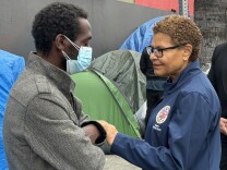 A woman with short hair (right) wearing a blue jacket with the L.A. city seal on it reaches out and holds the arm of a man (left) who's closing his eyes and wearing a mask, in front of tents in the background.